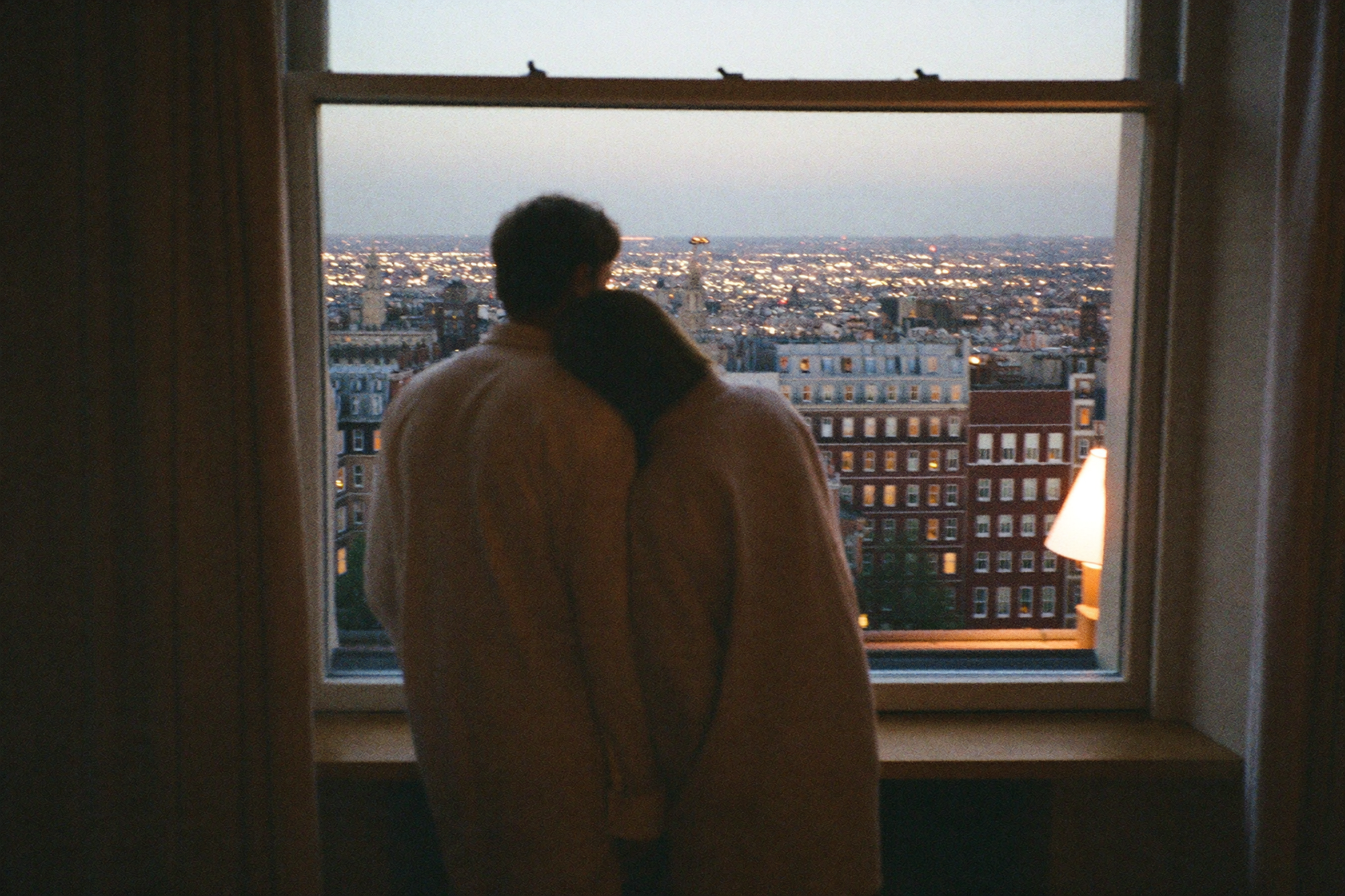 A couple at a window at dusk with the lights of London beyond the glass