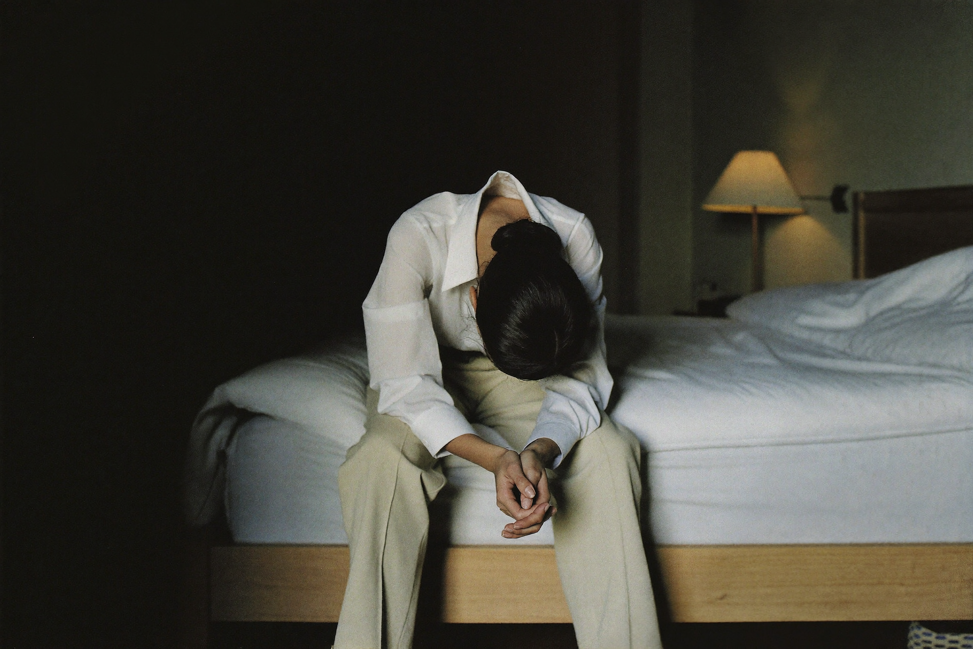 A woman sitting on the edge of her bed at night, shoulders dropped after a hard day