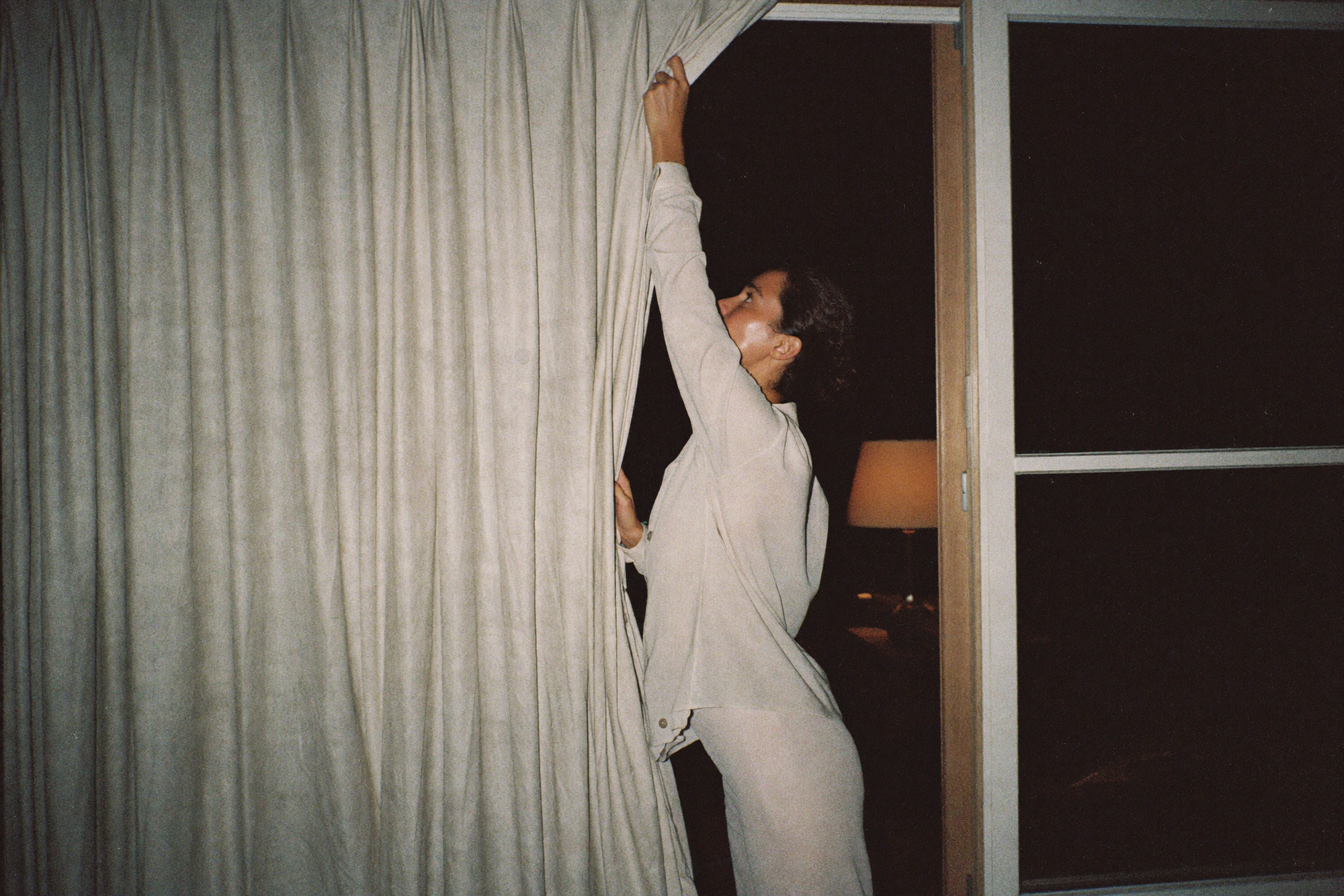 A woman drawing a heavy linen curtain closed across a bedroom window