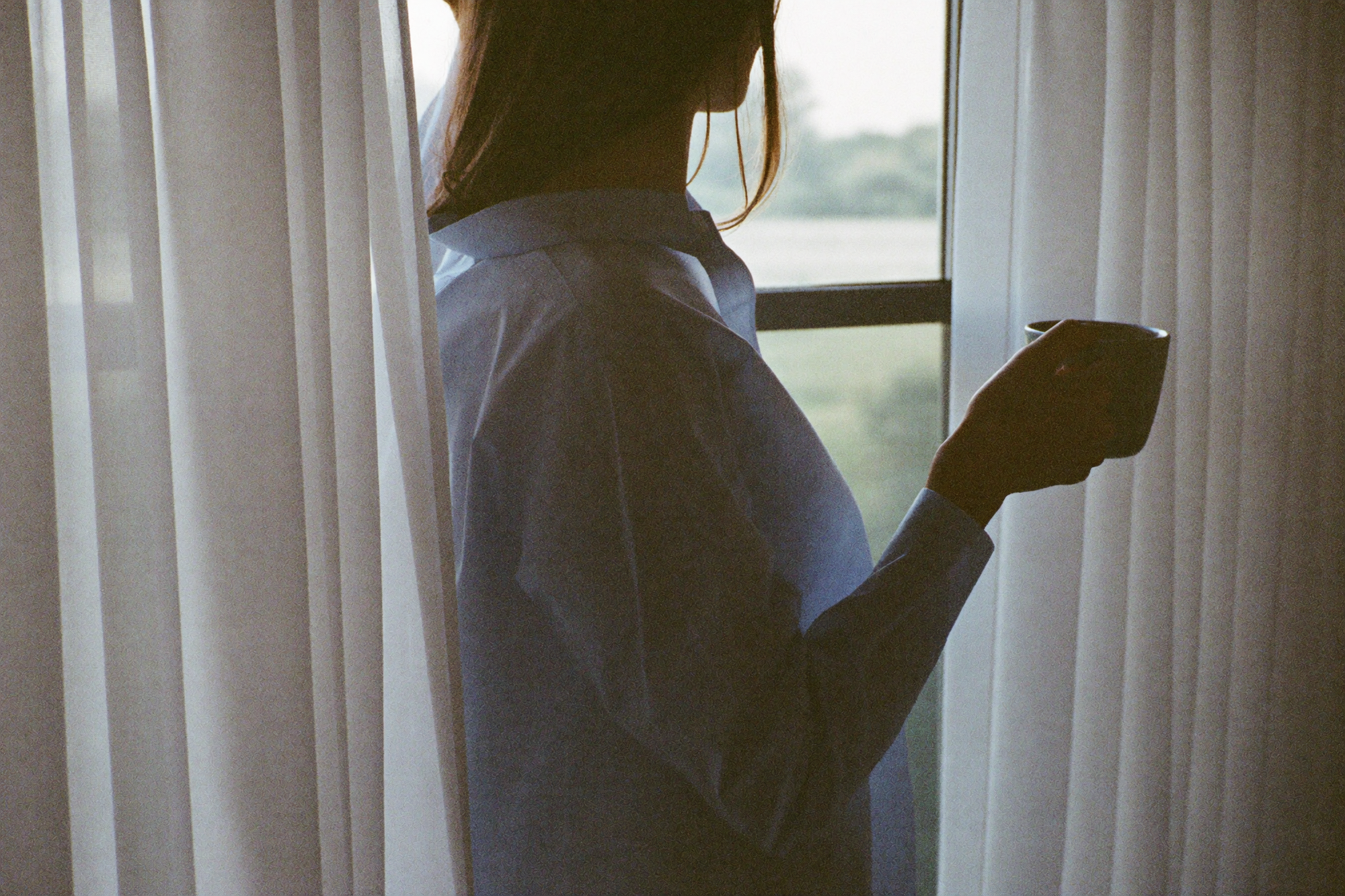 A woman at a bedroom window holding a mug in the flat light of early morning