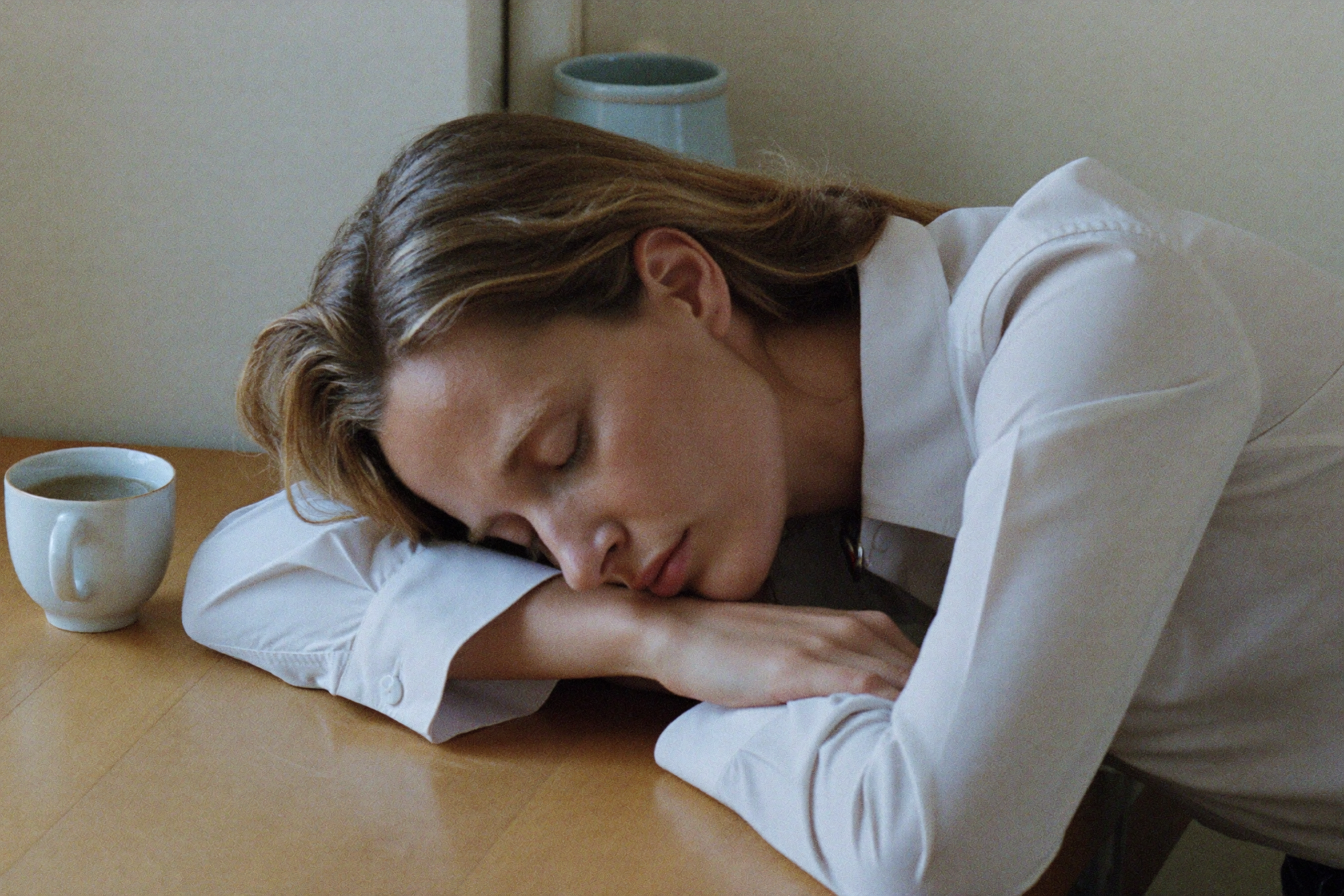 A woman asleep with her head on her arms at a table, beside an untouched cup