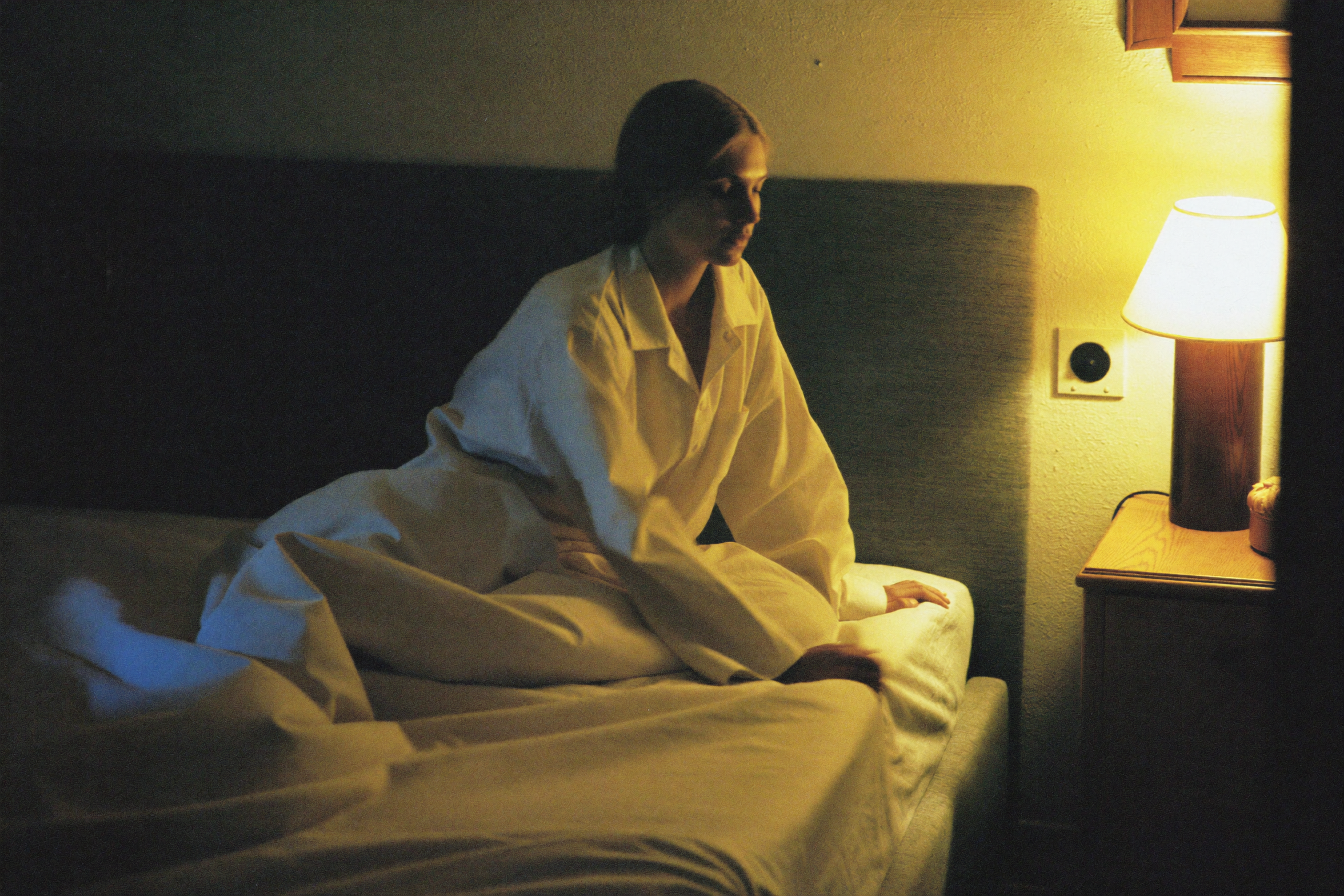 A woman in linen pyjamas turning down the corner of a made bed by lamplight