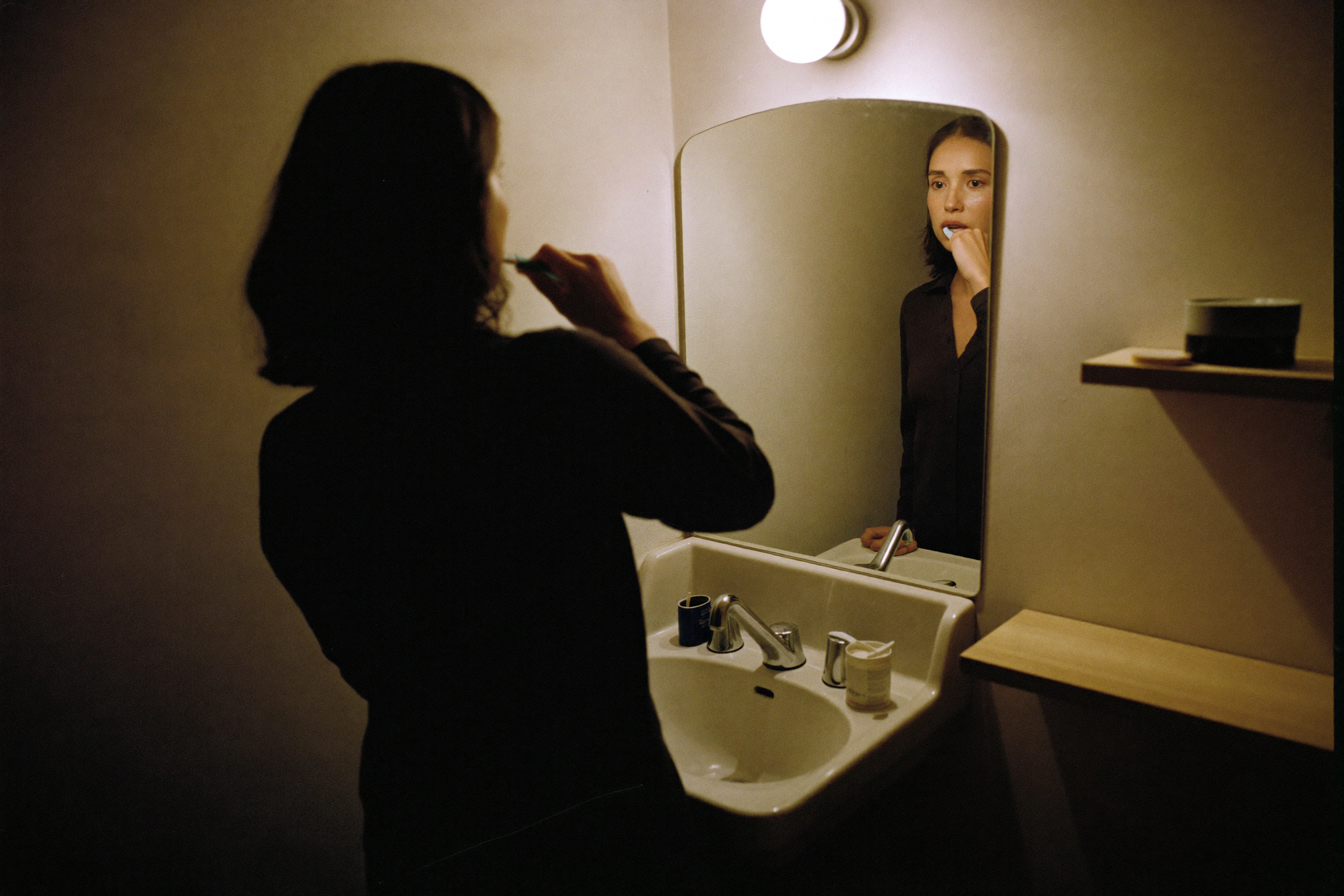 A woman brushing her teeth at a mirror very late at night