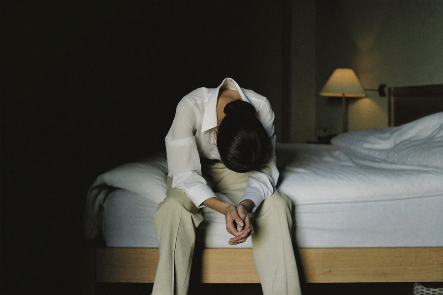 A woman sitting on the edge of her bed at night, shoulders dropped after a hard day