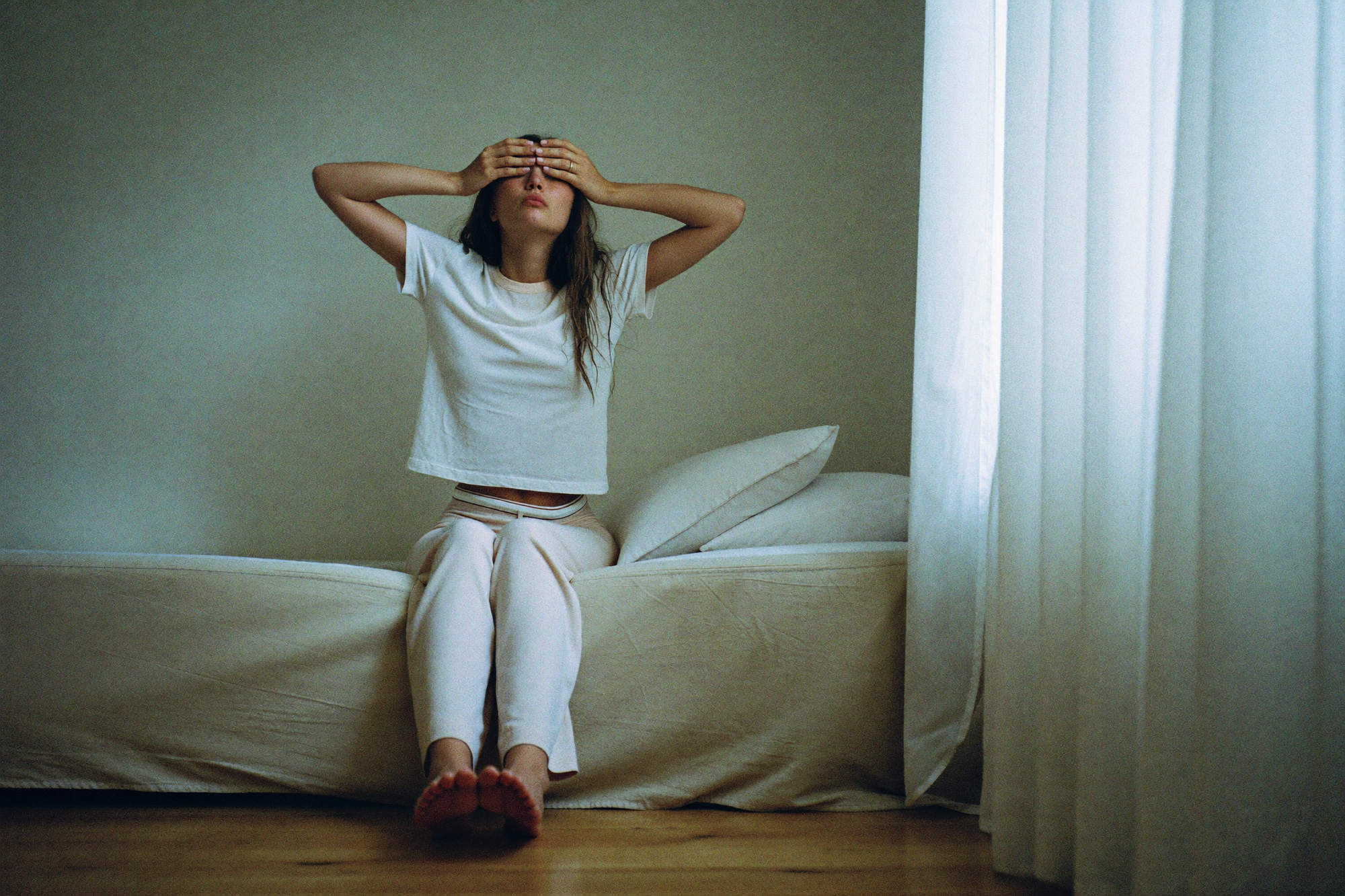 A woman on the edge of the bed pressing both hands into her eyes after waking