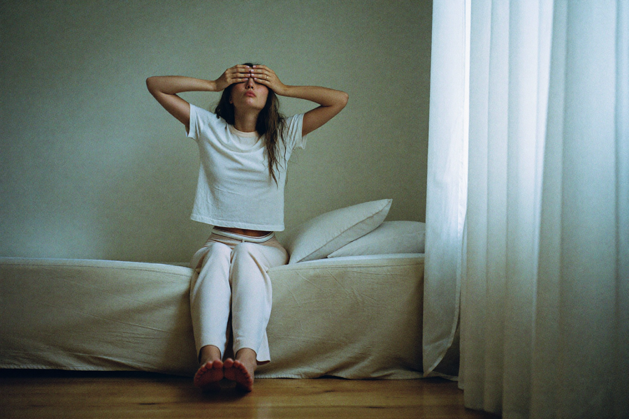 A woman on the edge of the bed pressing both hands into her eyes after waking
