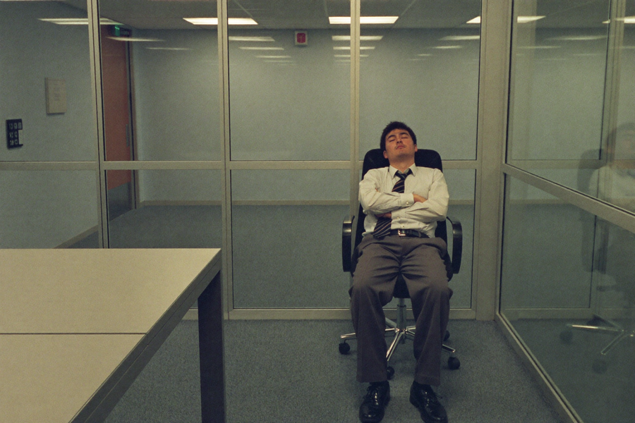 A man asleep upright in a reclined office chair in an empty meeting room
