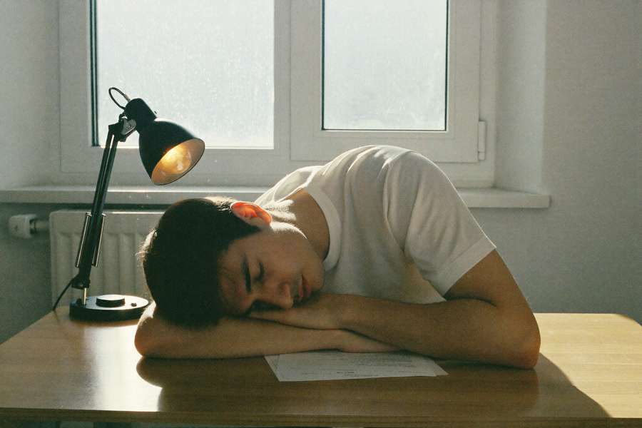 A man asleep at his desk as cold dawn rises behind him, the lamp still on